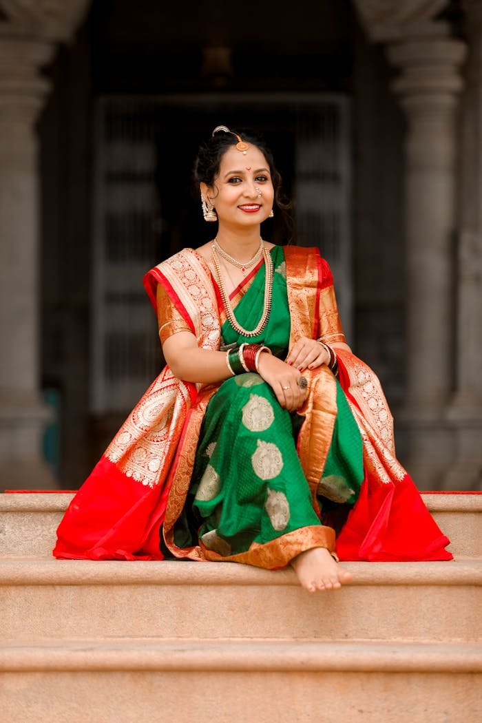 Smiling woman in a colorful saree sitting on steps outdoors.