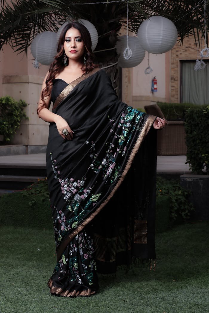 An elegant woman in a black saree with floral designs standing on grass with lanterns.