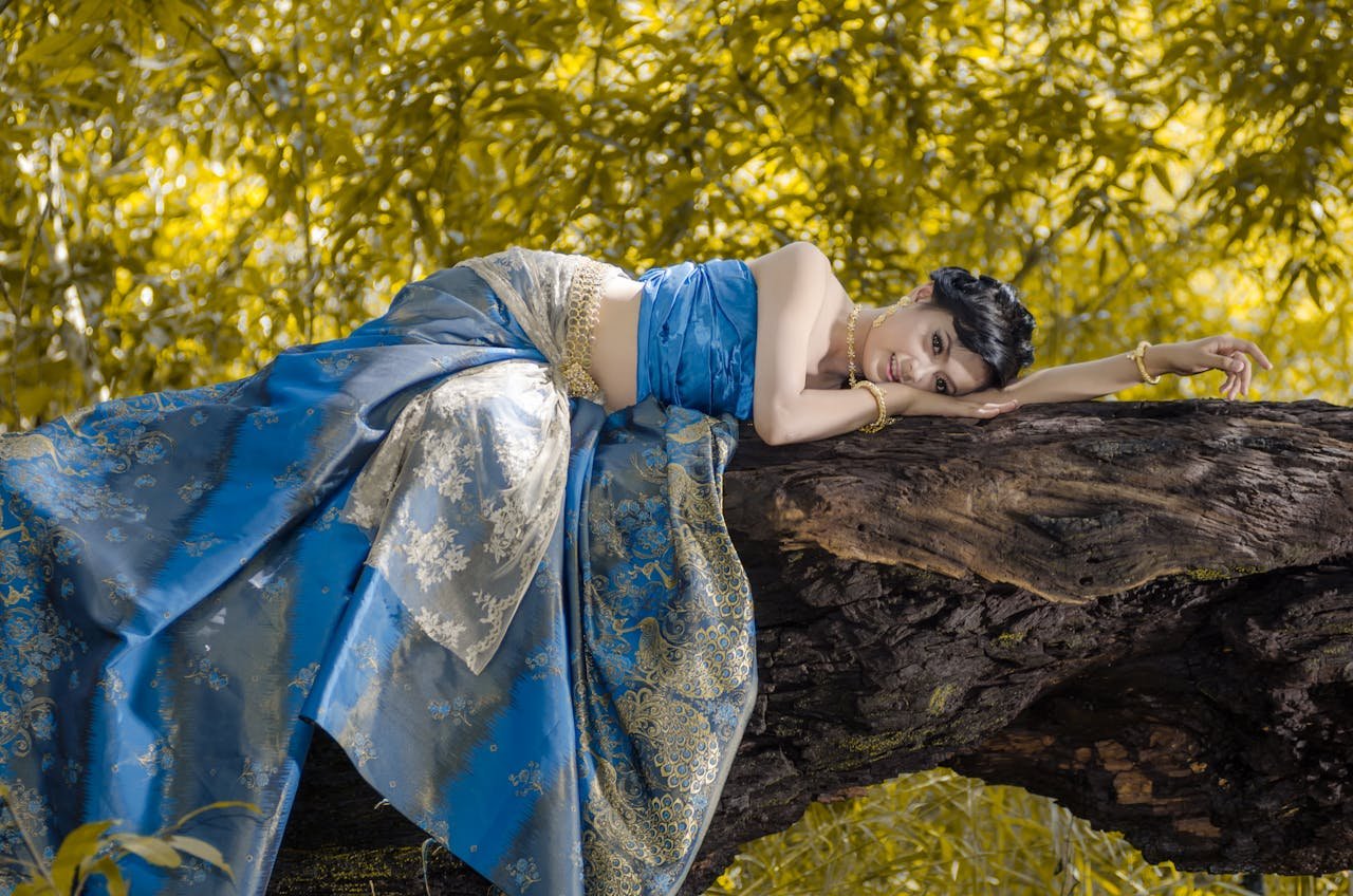 A woman in a traditional blue saree gracefully resting on a tree trunk with a serene backdrop.